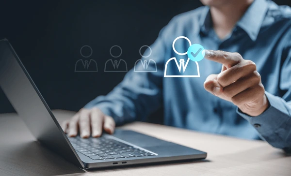 A person sitting at a desk uses a laptop while touching a virtual screen with user icons, selecting one icon marked with a blue tick—perfectly illustrating how to contact Golden Hire Recruitment.